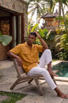 Man in a yellow linen short sleeve shirt and white linen pants sitting on a wooden chair by a pool with tropical balinese plants in the resort.