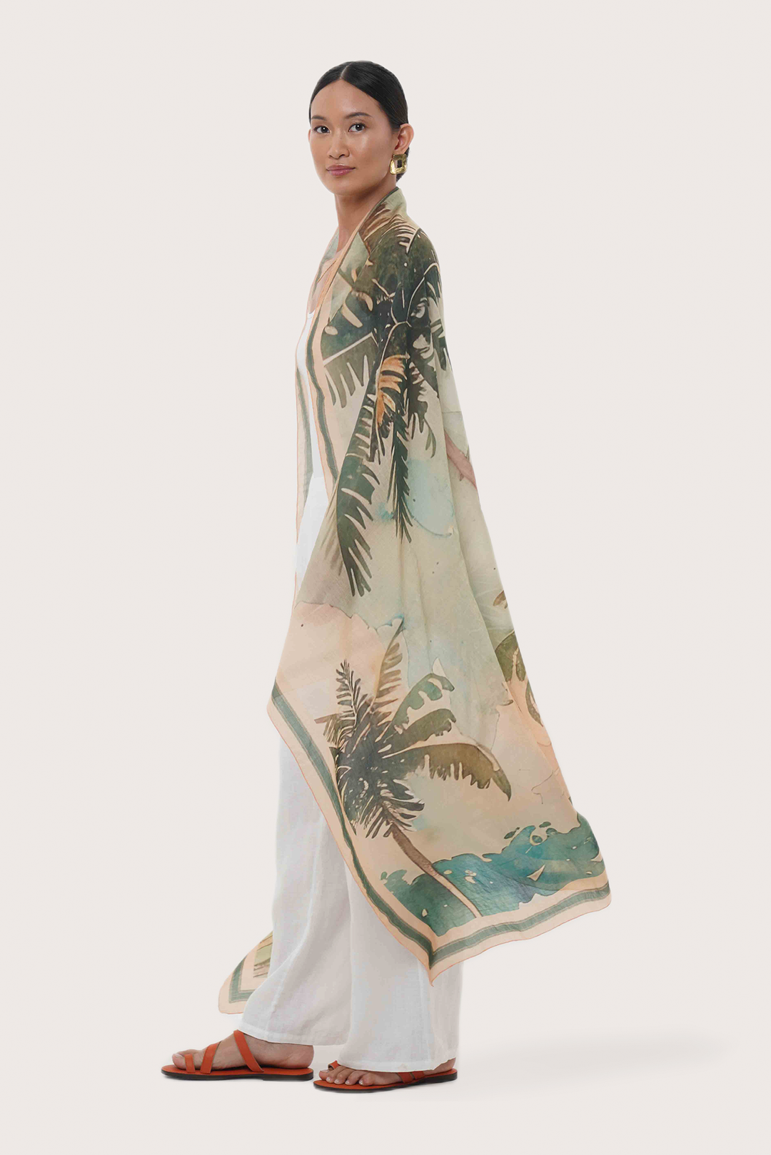 A woman modeling a lightweight draped sarong with a tropical palm tree print in green, beige, and teal, worn over white wide-leg pants and brown sandals against a light gray studio background.