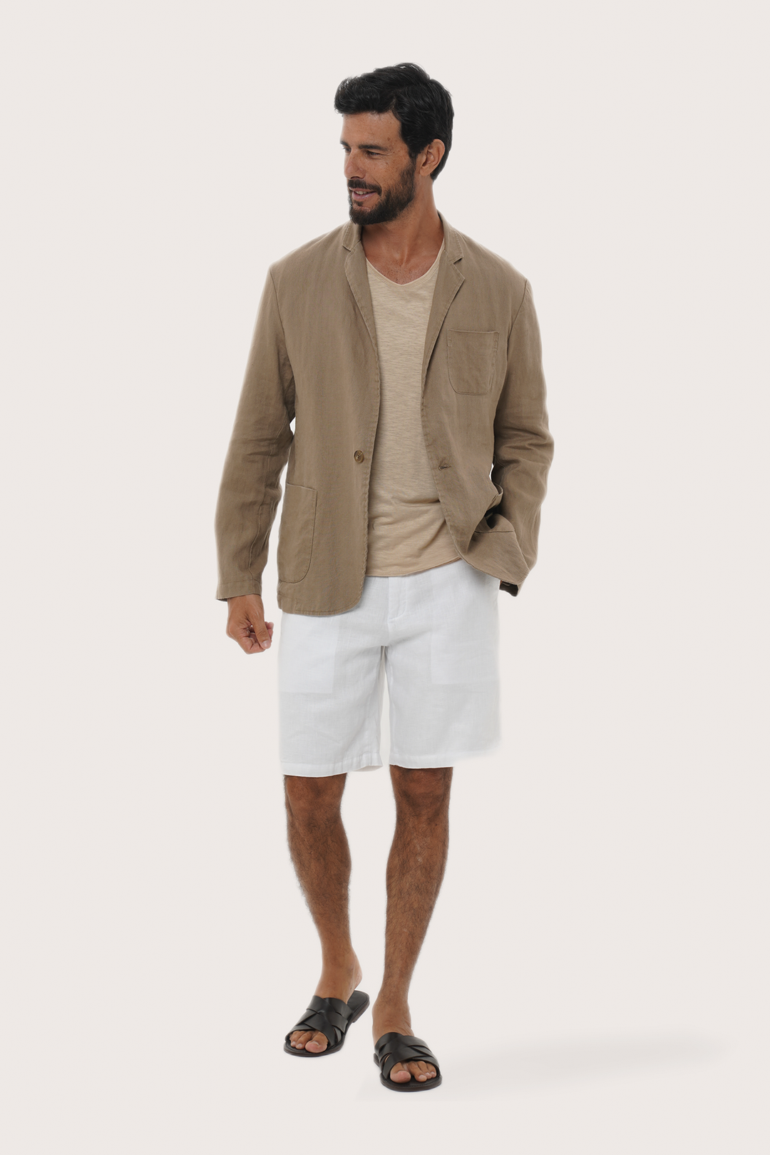 A man modeling a By the sea light brown linen-blend blazer over a beige t-shirt, white linen shorts, and black slide sandals, set against a solid off-white background.