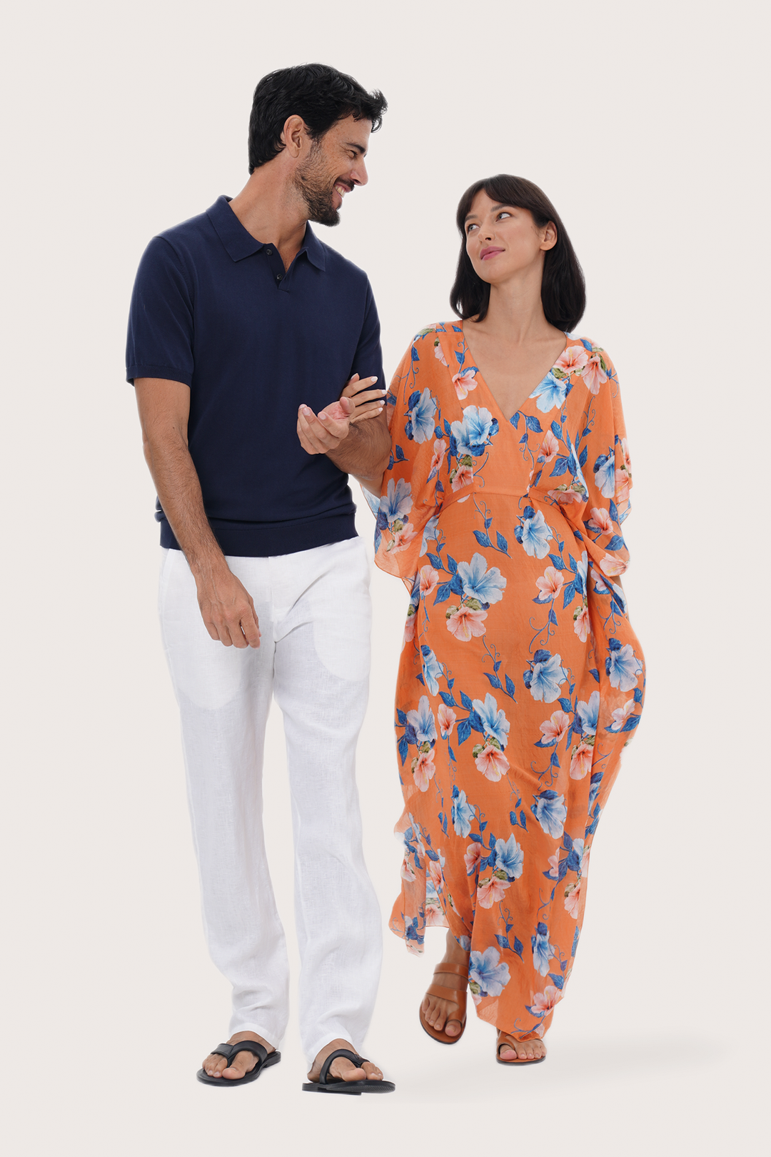 A man and a woman standing together against a plain light background. The woman is wearing a flowing orange floral dress with blue and white flowers, and the man is wearing a By the sea navy knit polo shirt, white linen pants, and leather sandals.