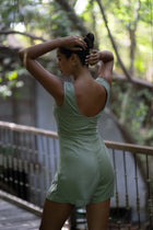 A woman wearing a sage green sleeveless romper stands on a wooden deck in a lush, green outdoor setting in bali
