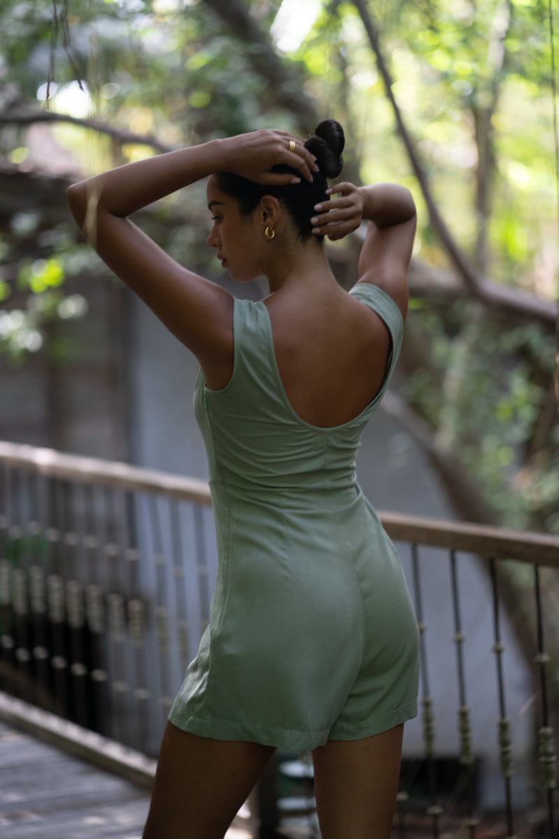 A woman wearing a sage green sleeveless romper stands on a wooden deck in a lush, green outdoor setting in bali