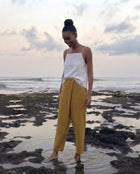 A woman stands barefoot in shallow tide pools on a rocky beach in bali, wearing a white sleeveless top and mustard yellow wide-leg pants.