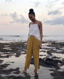 A woman stands barefoot in shallow tide pools on a rocky beach in bali, wearing a white sleeveless top and mustard yellow wide-leg pants.