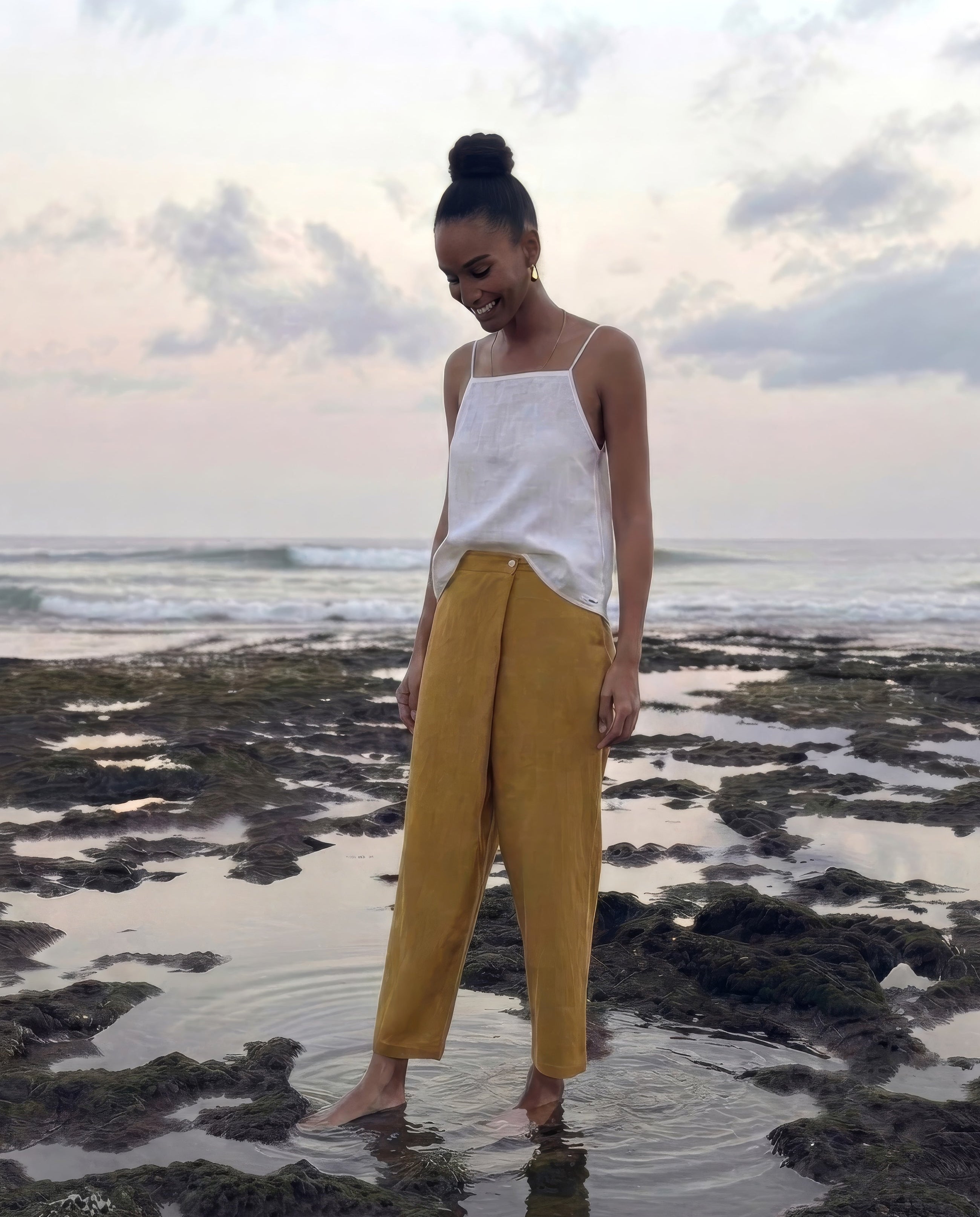 A woman stands barefoot in shallow tide pools on a rocky beach in bali, wearing a white sleeveless top and mustard yellow wide-leg pants.