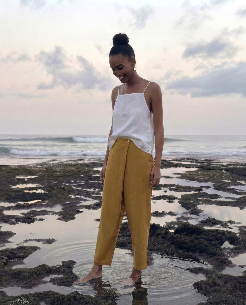 A woman stands barefoot in shallow tide pools on a rocky beach in bali, wearing a white sleeveless top and mustard yellow wide-leg pants.